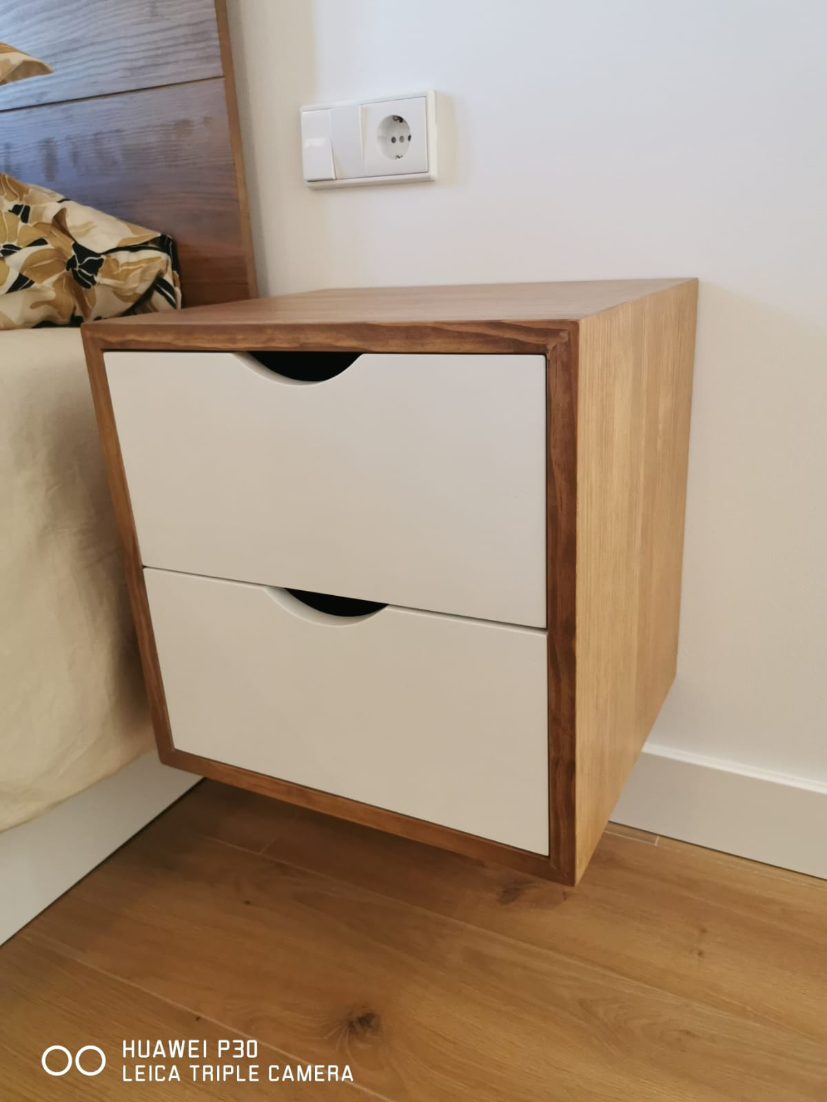 Mesita de noche con estructura de madera maciza y cajones de frente blanco junto al cabecero
